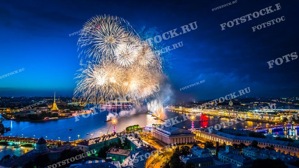 На фотографии представлен праздничный салют в Санкт-Петербурге, сделанный с высоты птичьего полета во время вечернего времени. Салют раскрывается над Невой, освещая водную гладь и архитектурные достопримечательности города, такие как Зимний дворец и Петропавловская крепость. Эта фотография может быть использована в материалах о праздниках и культурных мероприятиях, для промо-материалов туристических агентств, а также в публикациях, посвященных празднованию крупных городских событий.
